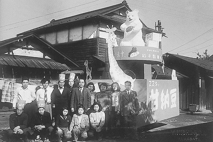 1953年当時の鈴木製菓（でん六へ社名変更前）と、「スズキの甘納豆」をPRする山車の前にて。傳六社長（後列左から5番目）と傳四郎専務（後列左から3番目）、ご家族や従業員の皆さん（写真提供：でん六）
